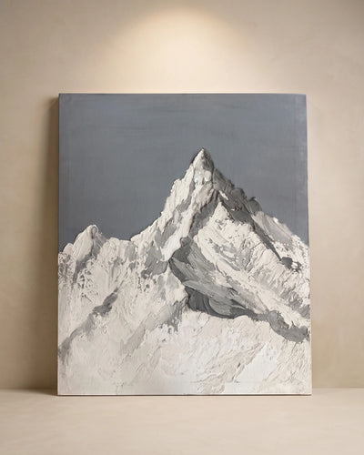 A square canvas painting of a snow-covered mountain peak, rendered in white, gray, and black brushstrokes against a muted gray sky, leaning against a beige wall under a spotlight.