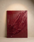 An abstract rectangular wall art piece in deep burgundy red, featuring textured, draped, and crinkled paint strokes, displayed against a neutral beige wall with a spotlight above.
