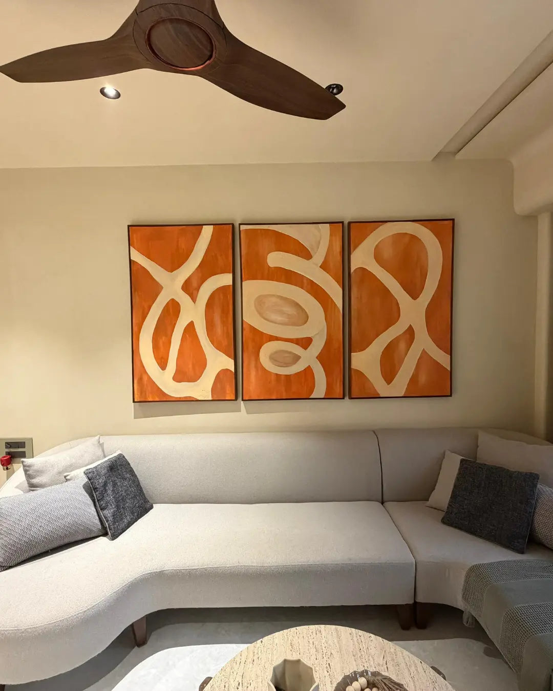 A 3-panel abstract canvas wall art set with orange and cream organic patterns, displayed above a modern curved light gray sectional sofa in a living room setting.
