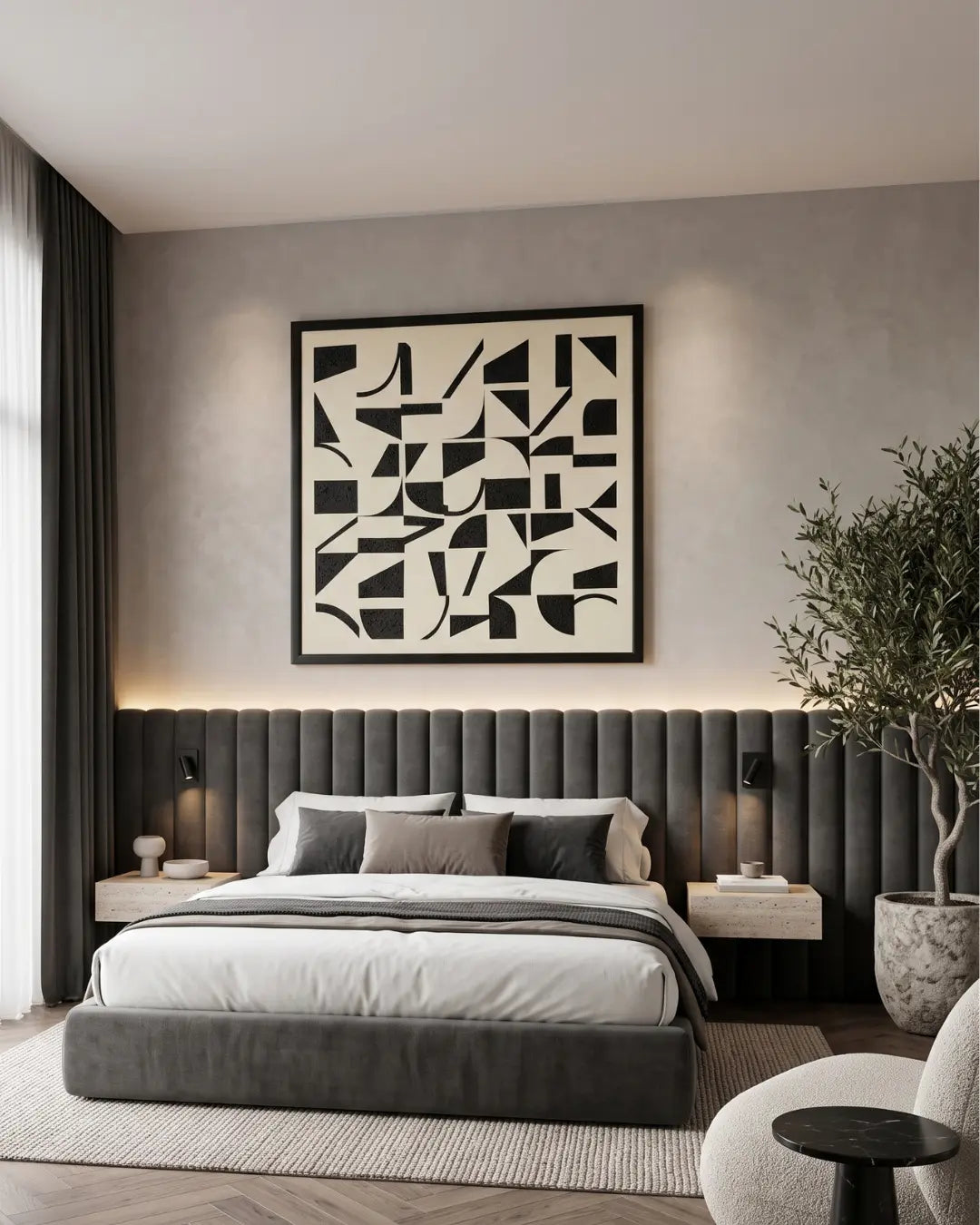 A modern bedroom featuring a low gray upholstered bed with a tall channel-tufted headboard, white bedding, bedside tables with lamps, a large black-and-white abstract wall art print, a potted tree, and a light gray textured wall.