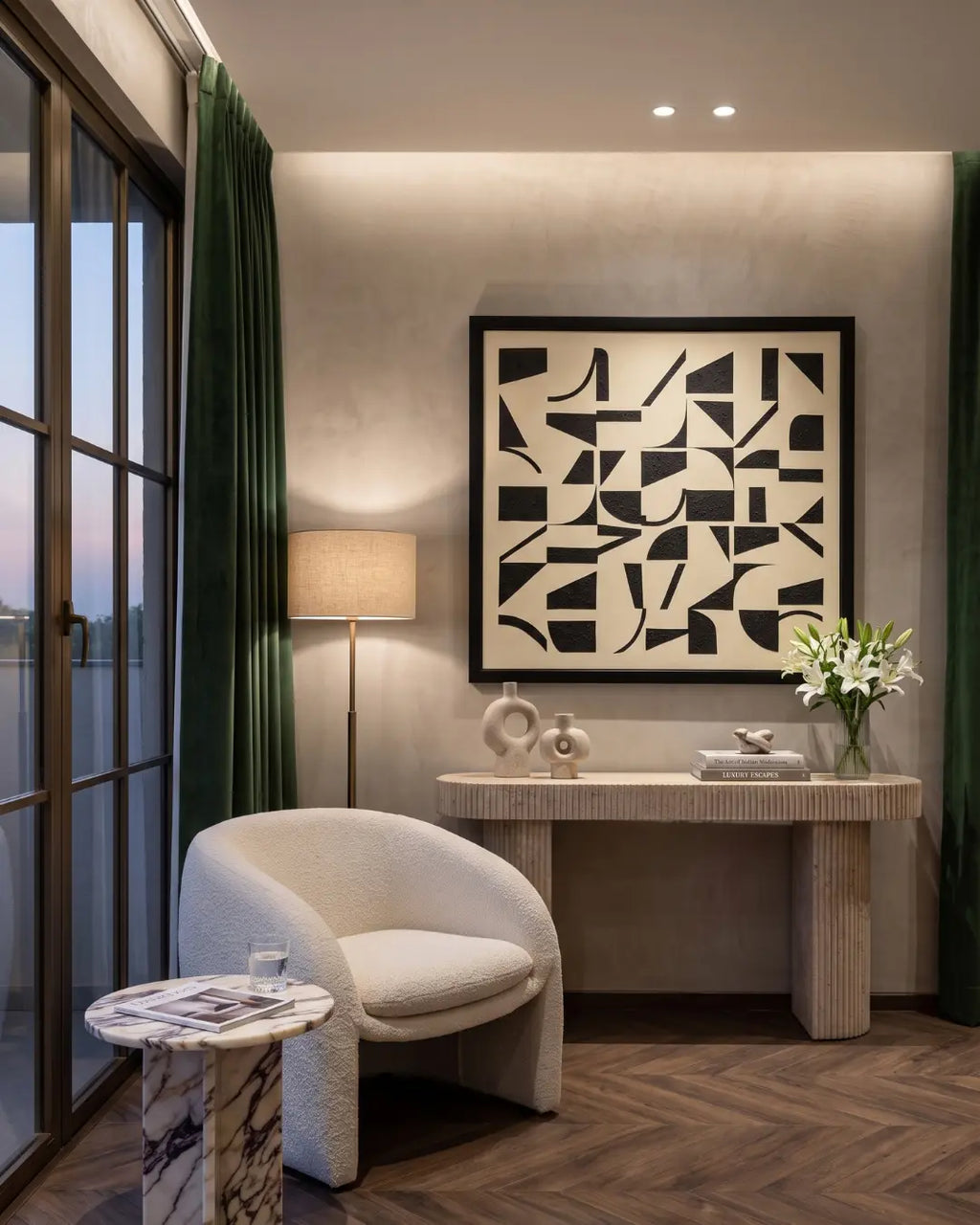 A modern interior room featuring a large black and white abstract geometric framed wall art piece hanging above a textured stone console table. The room includes a curved cream upholstered armchair, a small marble-topped side table with a glass, a floor lamp, green curtains, and a herringbone wood floor.