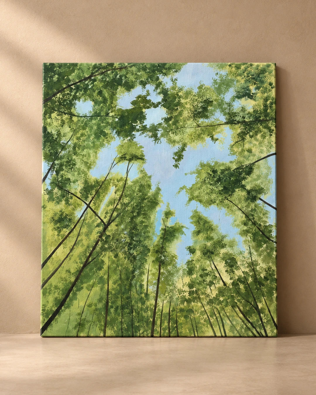 A square canvas painting of a forest canopy viewed from below, with green trees and leaves framing a light blue sky, displayed against a beige wall on a light wood surface.
