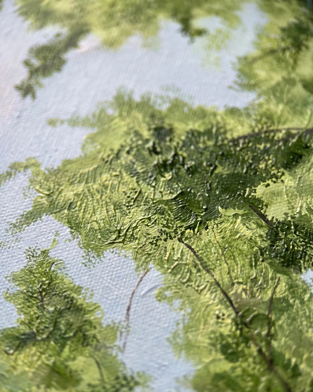 A close-up of a textured painting depicting green trees and foliage reflected in light blue water, with visible canvas texture and paint strokes.