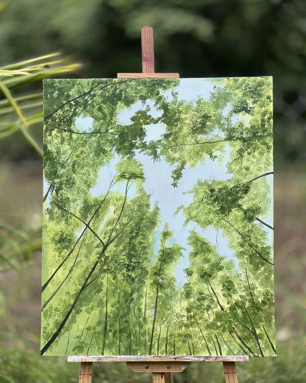 A canvas handmade painting of a forest canopy with green trees and a light blue sky, displayed on a wooden easel outdoors against a blurred green background.