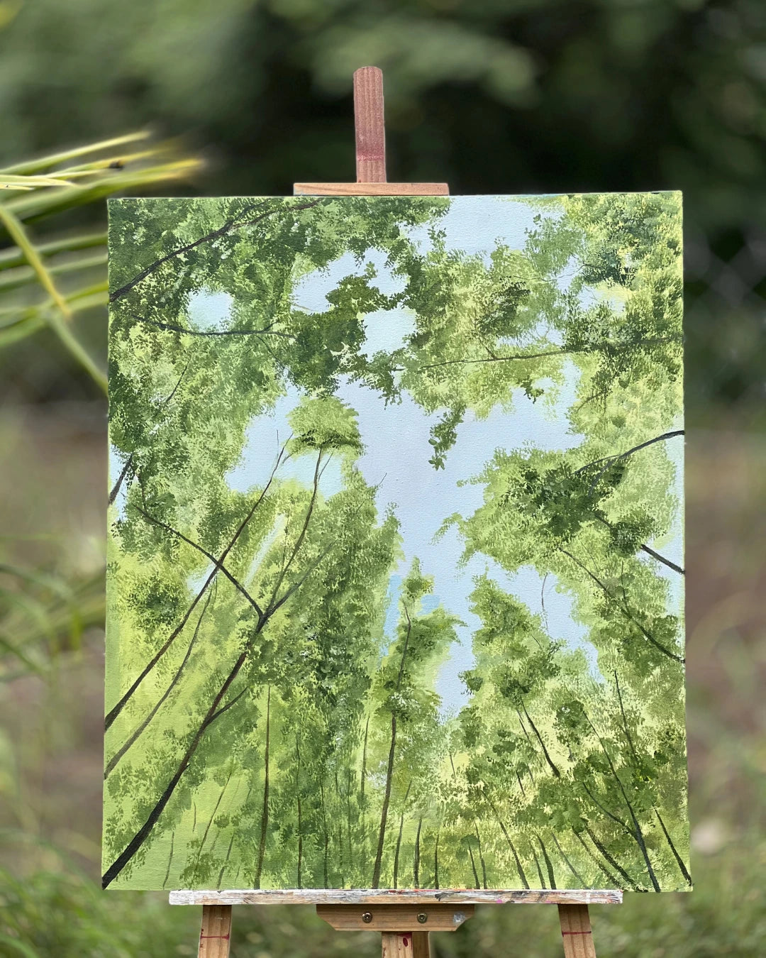 A canvas handmade painting of a forest canopy with green trees and a light blue sky, displayed on a wooden easel outdoors against a blurred green background.