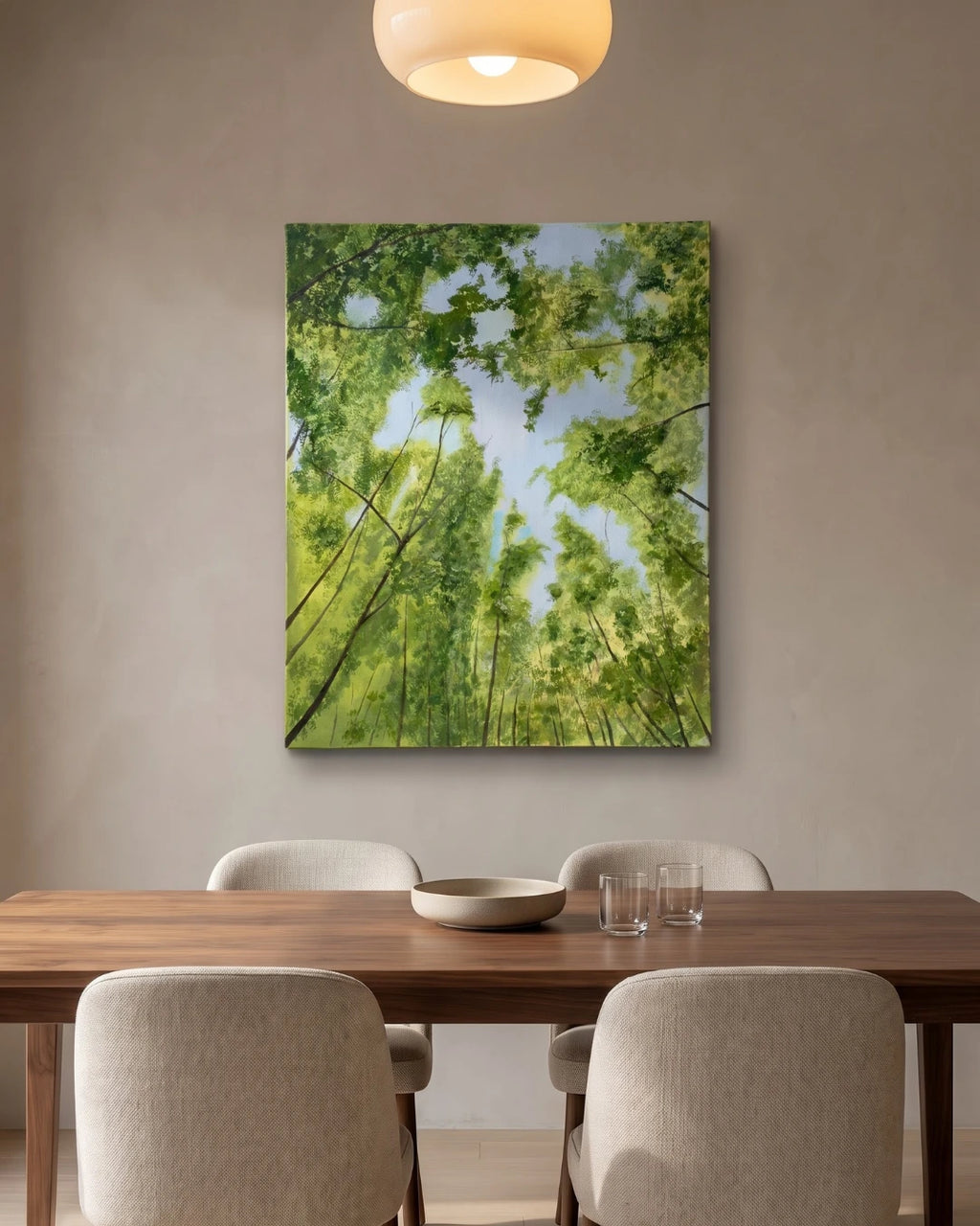 A vertical canvas wall handmade art depicting a lush green forest canopy looking up at the trees and blue sky, displayed on a neutral beige wall above a wooden dining table with four chairs.