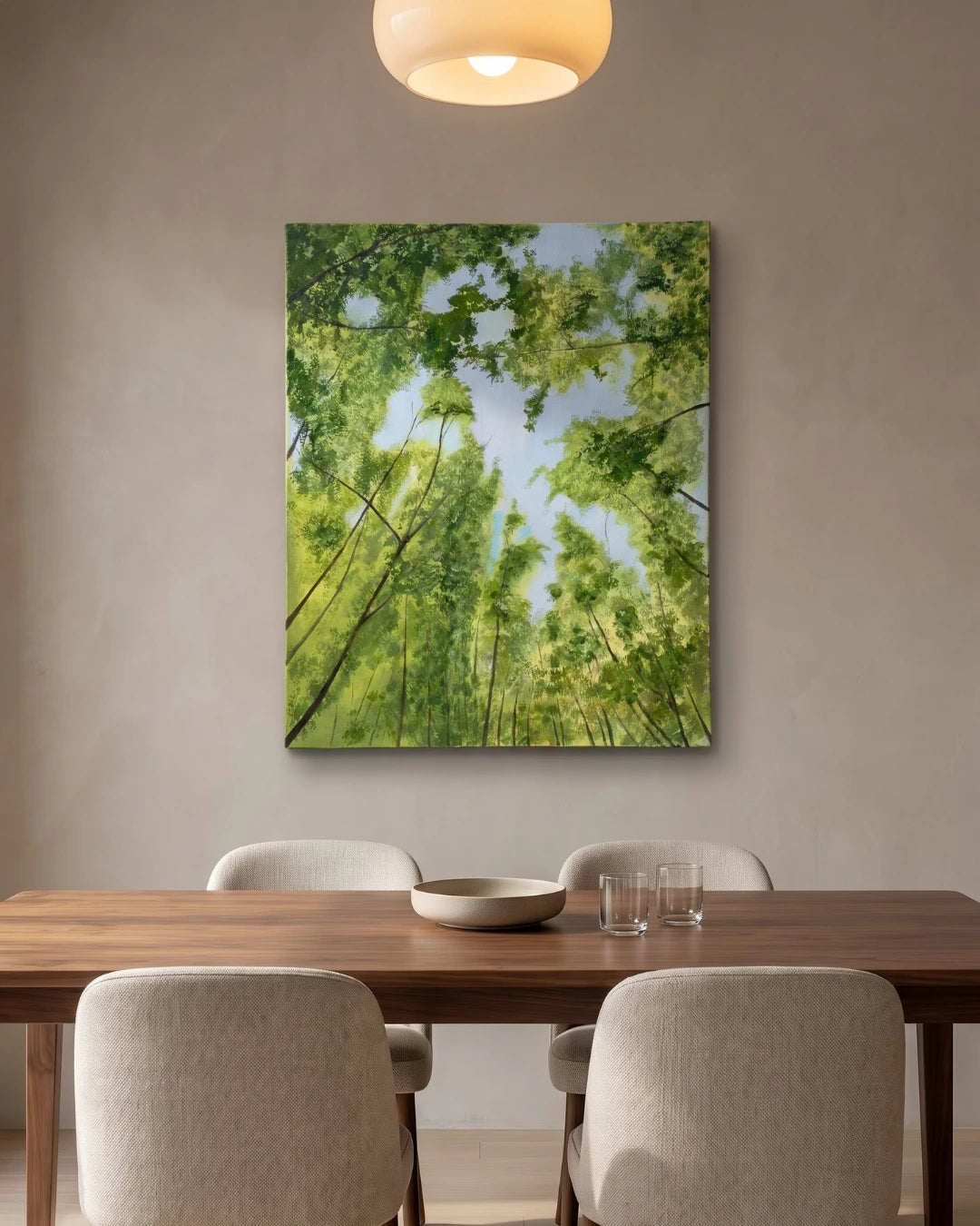 A vertical canvas wall handmade art depicting a lush green forest canopy looking up at the trees and blue sky, displayed on a neutral beige wall above a wooden dining table with four chairs.