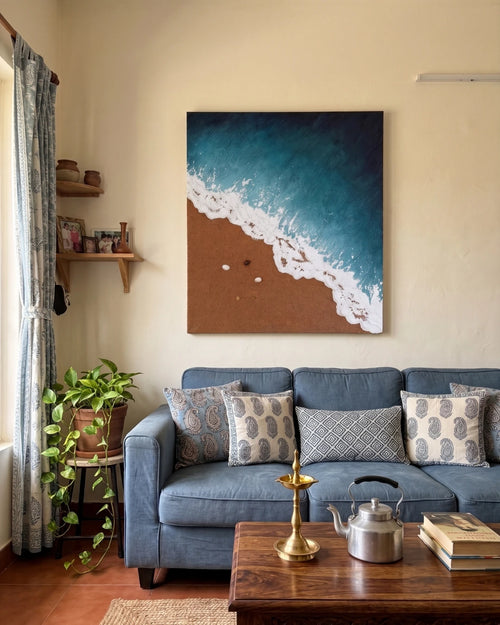 Textured ocean artwork with foamy white surf, sandy beach, and seashell details displayed above a cozy blue living room sofa.

