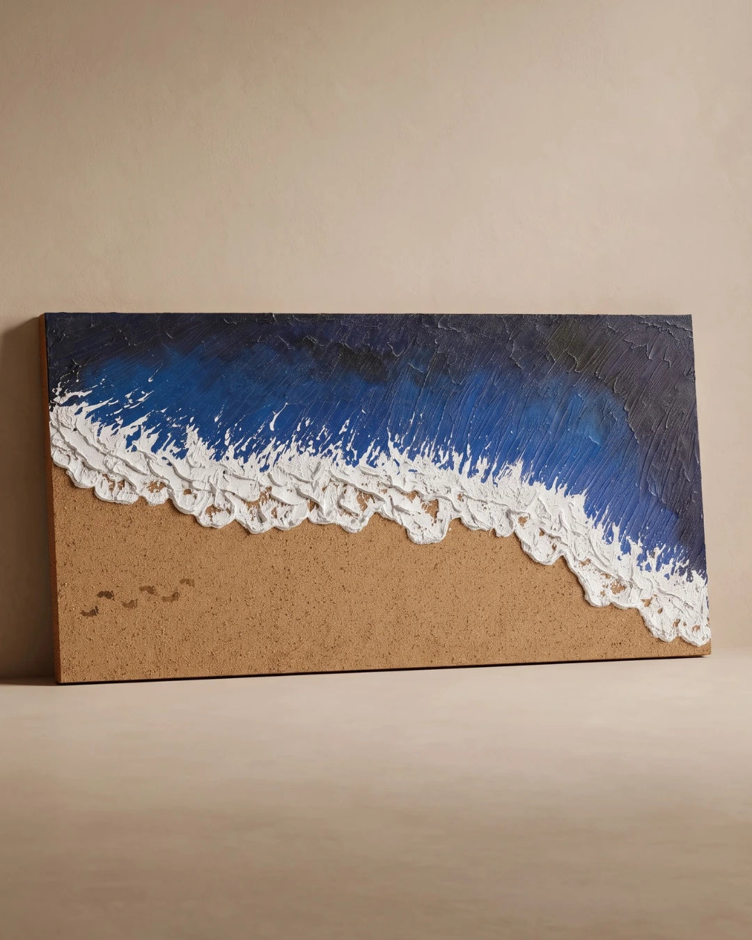 A rectangular canvas wall art piece depicting an ocean wave with textured blue and dark navy water, white foam, and a sandy beige shore, displayed against a plain beige wall.