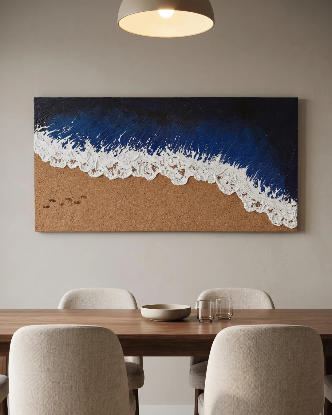 A large rectangular wall art piece showing a beach scene with deep blue ocean waves and white foam over tan sand, displayed above a wooden dining table with chairs in a modern interior.