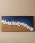 A rectangular wall art piece depicting an ocean wave on a sandy beach, with textured deep blue water, white foam, and tan sand, displayed against a warm beige wall.