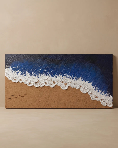 A rectangular wall art piece depicting an ocean wave on a sandy beach, with textured deep blue water, white foam, and tan sand, displayed against a warm beige wall.