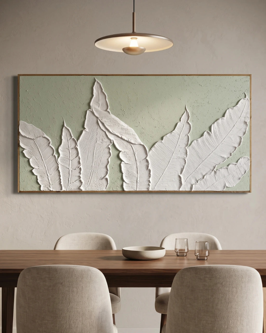 Large framed wall art featuring white 3D leaf-like forms on a pale sage green textured background, displayed above a wooden dining table with chairs in a modern interior.
