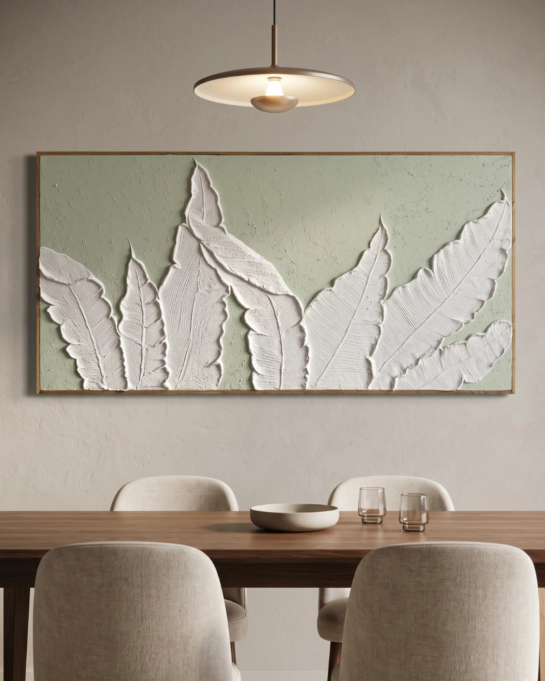 Large framed wall art featuring white 3D leaf-like forms on a pale sage green textured background, displayed above a wooden dining table with chairs in a modern interior.