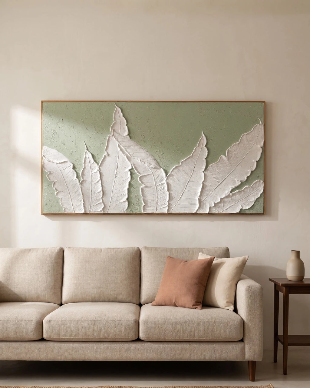A large framed wall art piece featuring white 3D leaf shapes on a muted sage green background, displayed above a light beige sofa in a neutral living room setting.