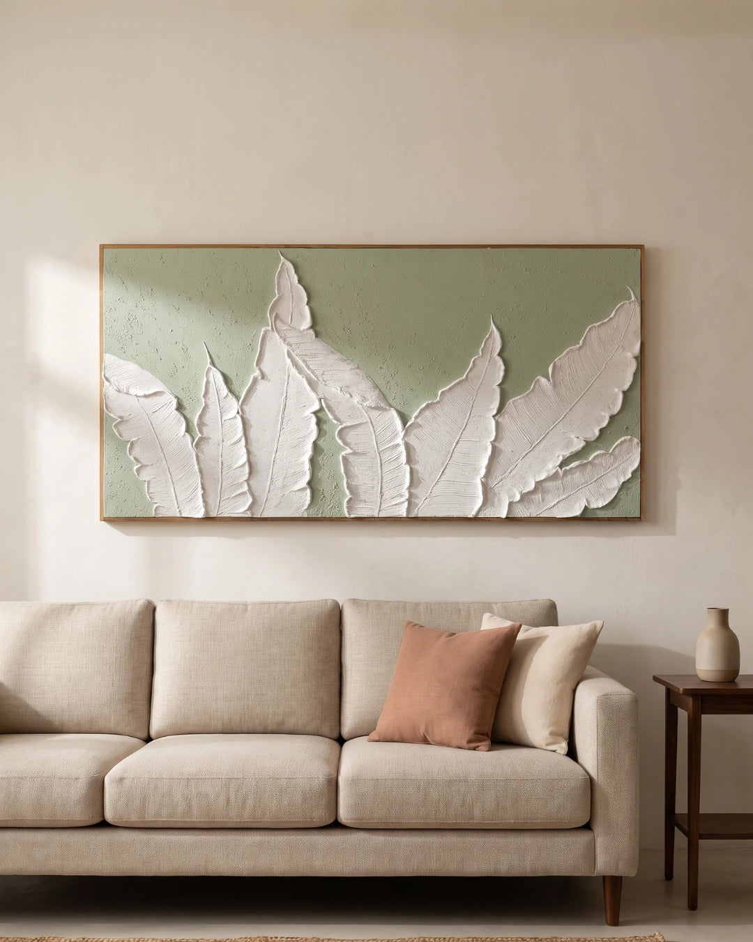 A large framed wall art piece featuring white 3D leaf shapes on a muted sage green background, displayed above a light beige sofa in a neutral living room setting.