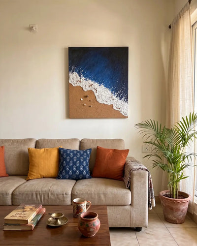 A square canvas wall art print depicting a beach scene with deep blue ocean waves crashing onto a sandy shore, hanging on a beige wall above a modern beige sofa with decorative throw pillows in a styled living room.