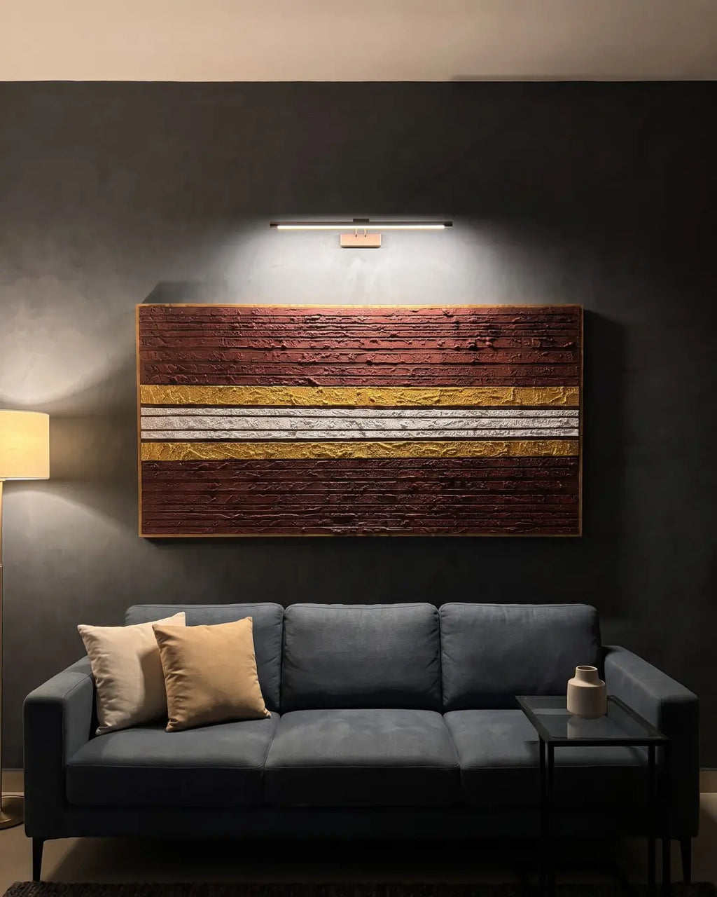 A rectangular textured wall art piece with horizontal bands of dark brown, golden yellow, and off-white, displayed on a dark gray wall above a blue-gray sofa in a modern living room setting.