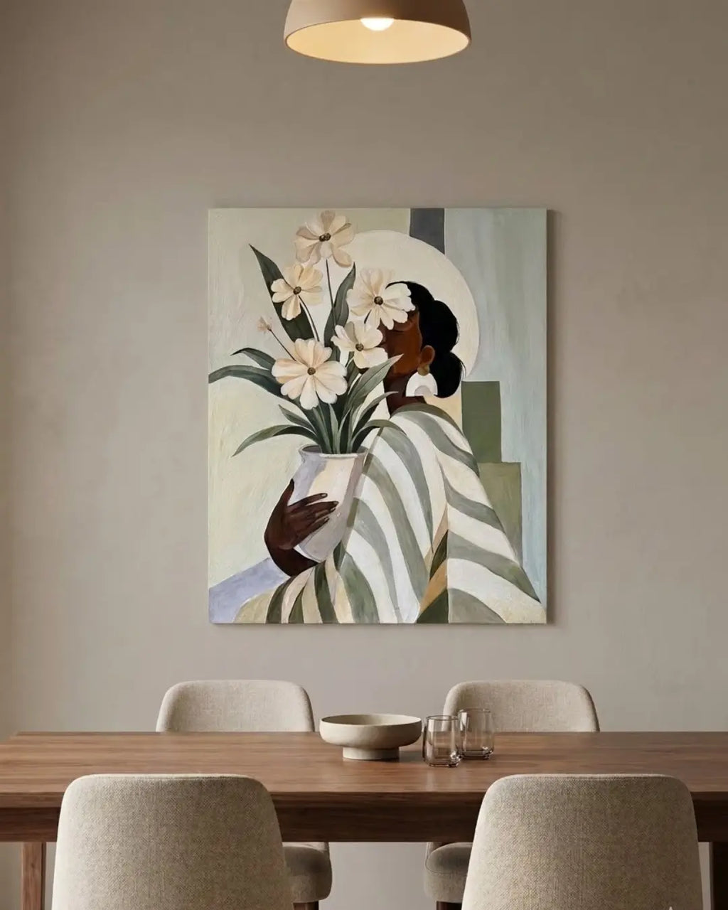 A framed wall handmade art depicting a stylized woman holding white flowers, displayed above a wooden dining table with neutral chairs in a modern interior setting.