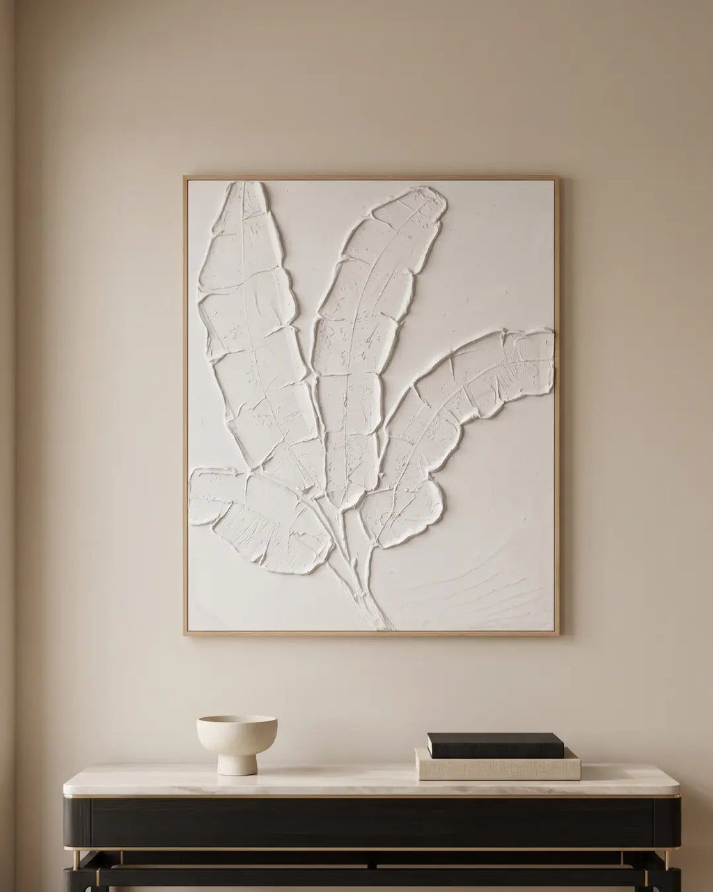Large framed wall art featuring a textured white leaf design, displayed on a beige wall above a console table with a bowl and books.